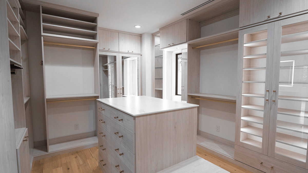 Image of a luxury custom walk-in closet featuring a closet island with drawers, display cabinet with glass doors, and multiple hanging sections for clothes.