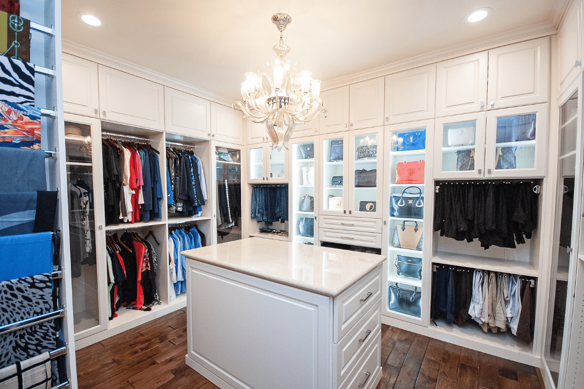 Image of a custom walk-in closet Houston, with built-in display cabinets for purses, a closet island, and chandelier.